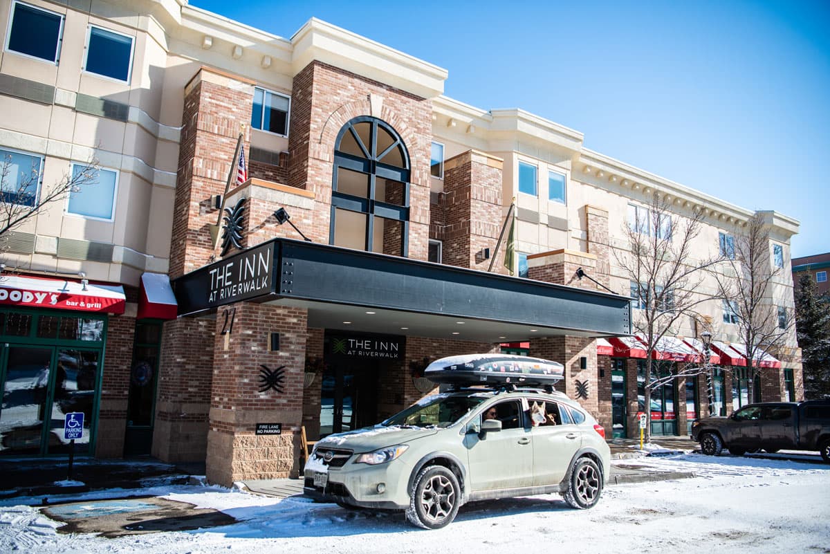 An outside photograph of the brick exterior of The Inn at Riverwalk in Edwards, Colorado. On the left and right sides of the exterior are more business and buildings on a quaint street in Edwards. On the curb is a light green Subaru covered in a light layer of snow. Two dogs are sticking their heads out the window and someone is in the front seat. The sky is blue above the building.