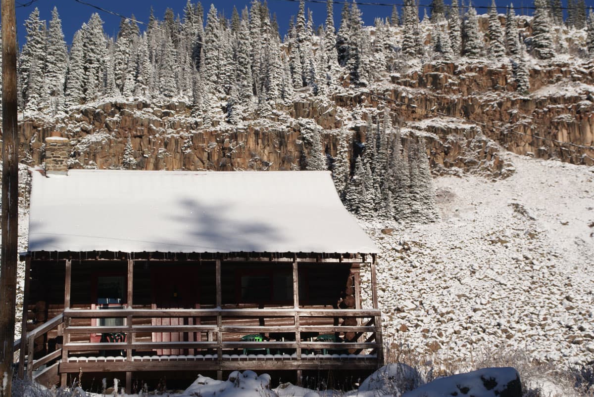 A rustic cabin at Grand Mesa Lodge is dusted with snow. The rugged landscape behind it is snowy, too, with a bright blue sky behind it.