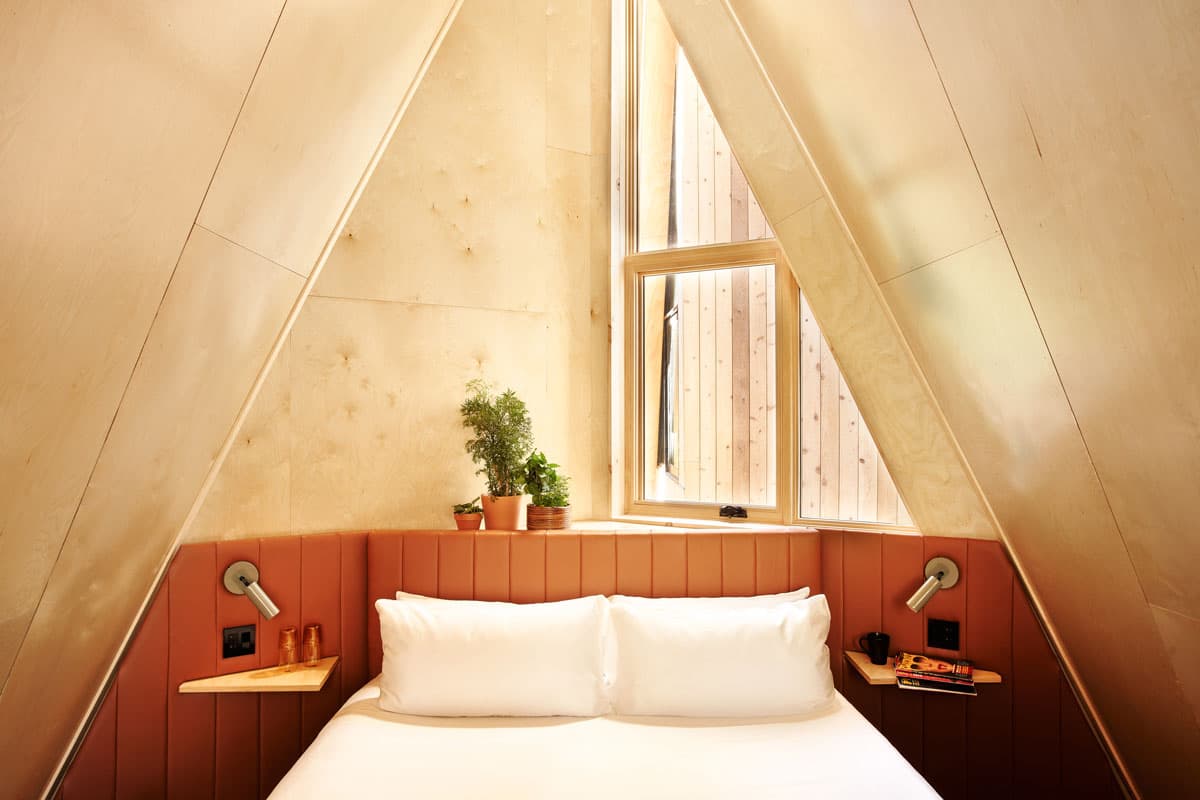 A comfy bed covered in white bedding sits under a triangular roof with white interior walls at A-Frame Club.