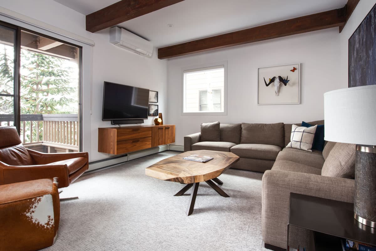 A condo unit at The Gant in Aspen features dark-wood ceiling beams, a plush tawny couch and a decorative coffee table