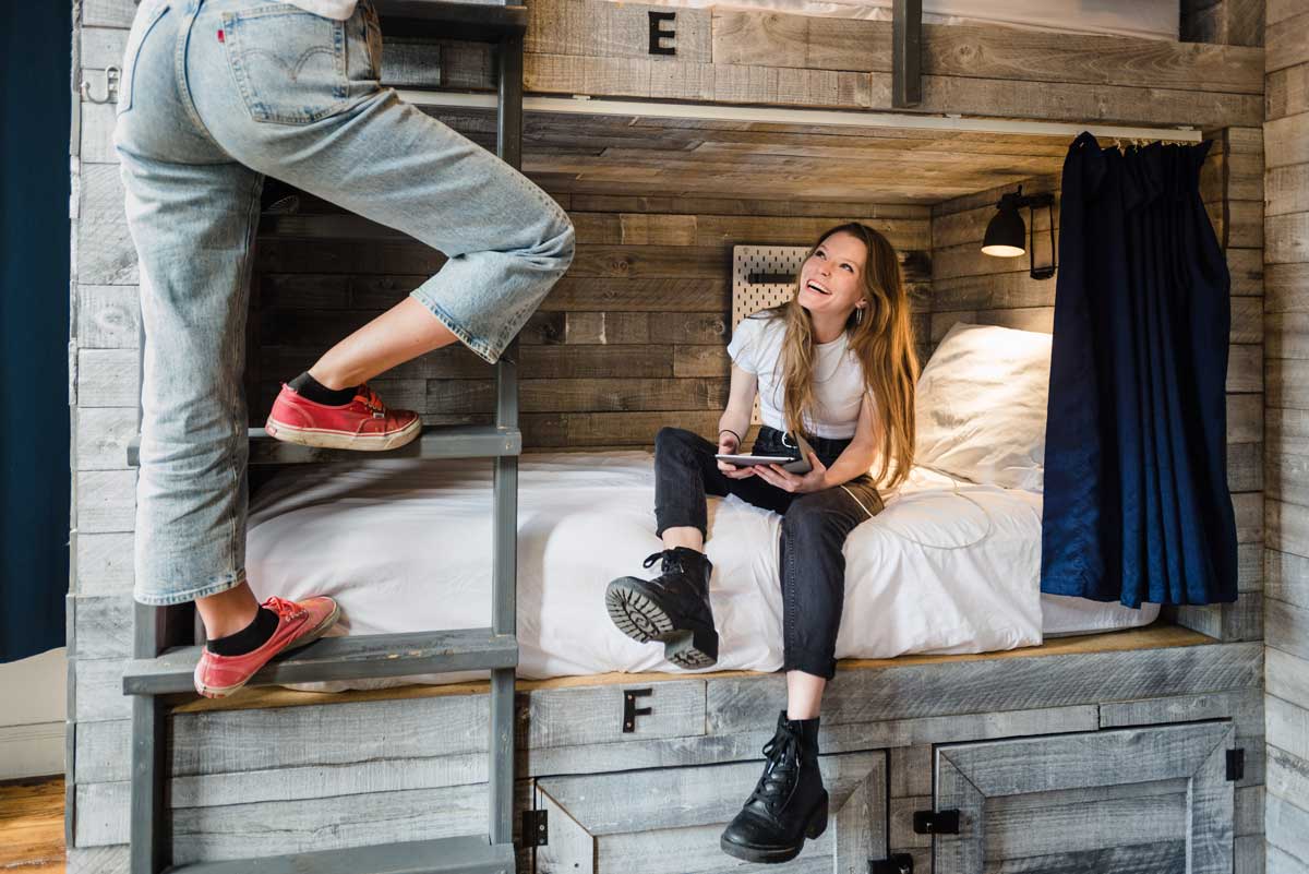 Girl in black jeans and boots sits on comfy bunk bed while chatting with another person who is climbing to the top bunk wearing jeans and red sneakers