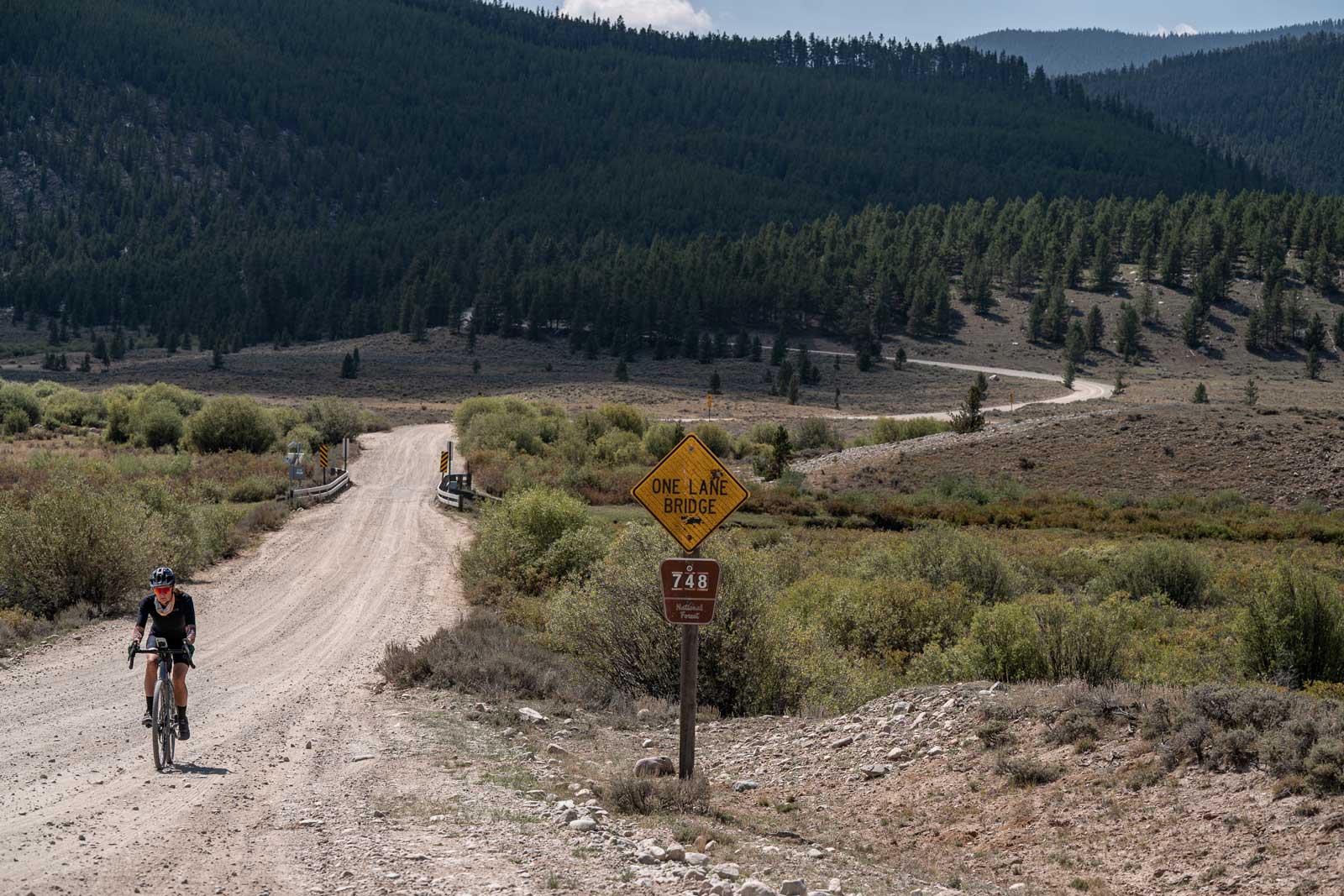 A gravel biker rides across a bridge next to a National Forest service marker near Crested Butte