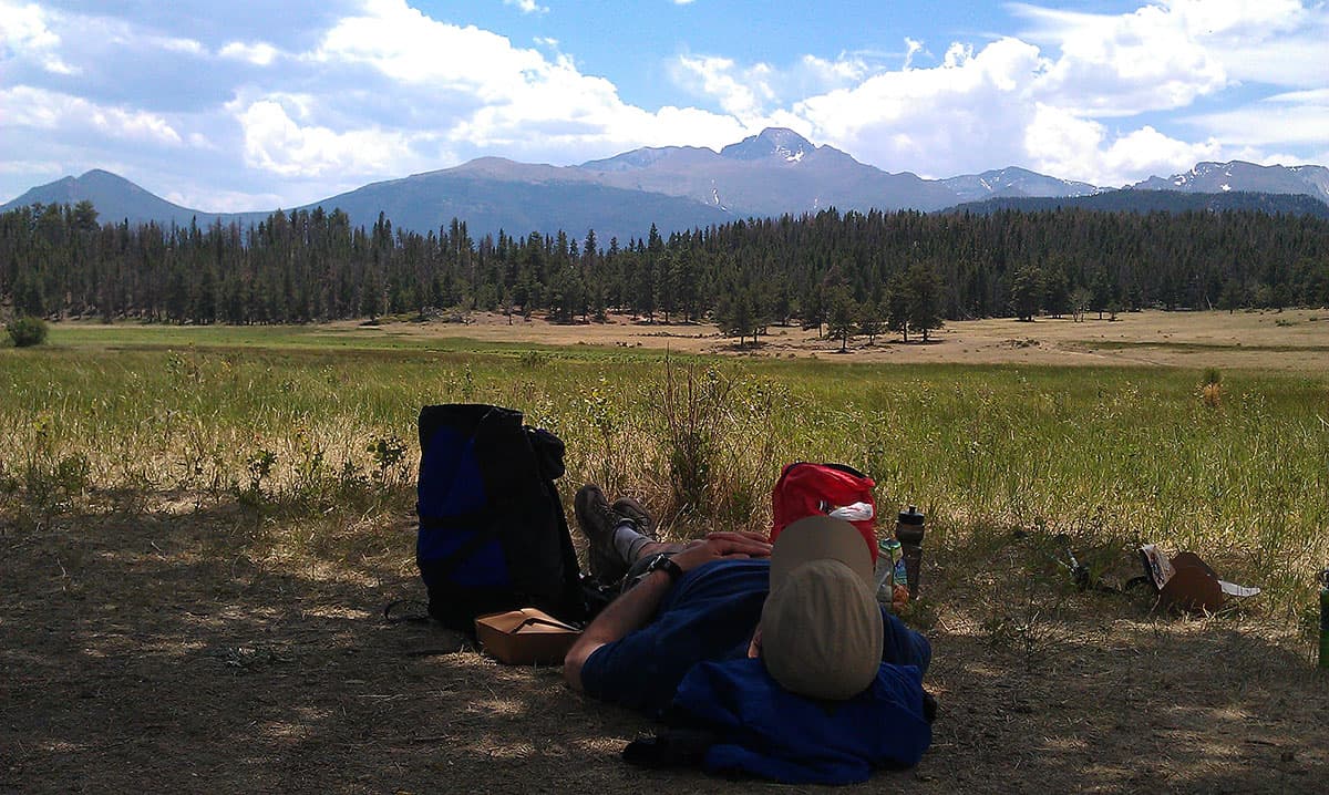 Man laying on the ground in the shade wearing a tan hat with mountain peaks and forest trees ahead of him