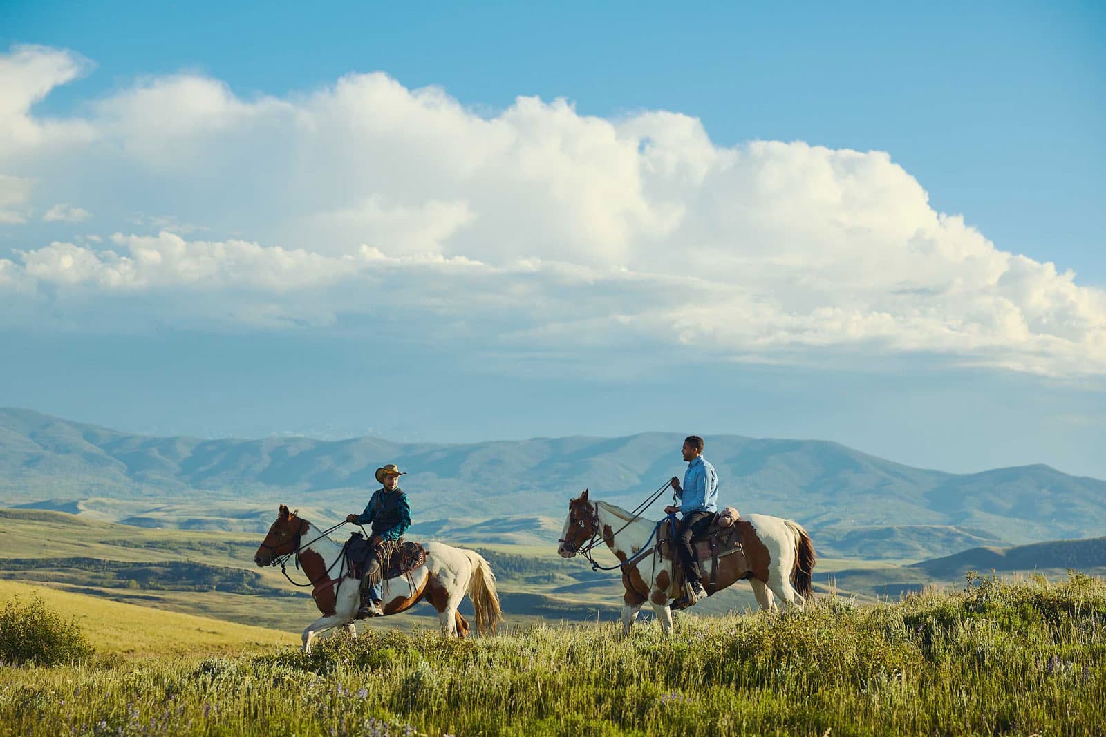 Two people ride horses through a grassy field with rolling green hills in the background