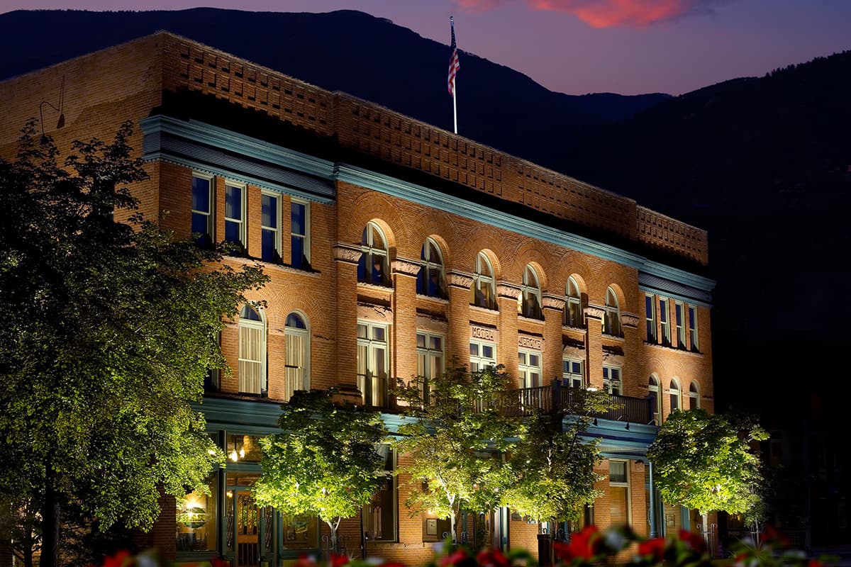 The red-brick facade of the historical Hotel Jerome is lit with lights. Behind it, the sun sinks behind the mountains creating pink clouds in the Colorado sky.