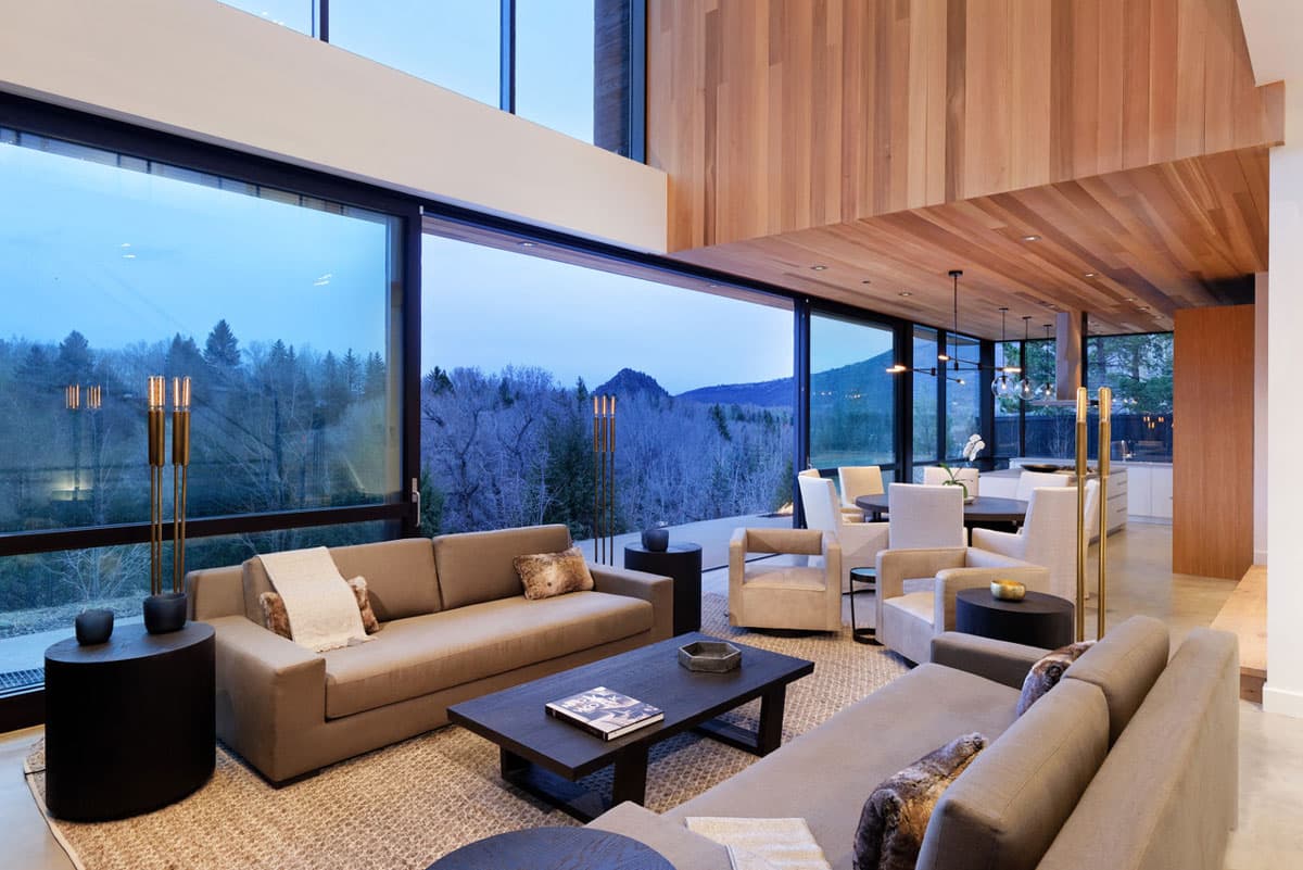 Great room with modern loungers and living room light fixtures, with a wide window behind revealing winter snow and forest nearing dusk in Aspen, Colorado.