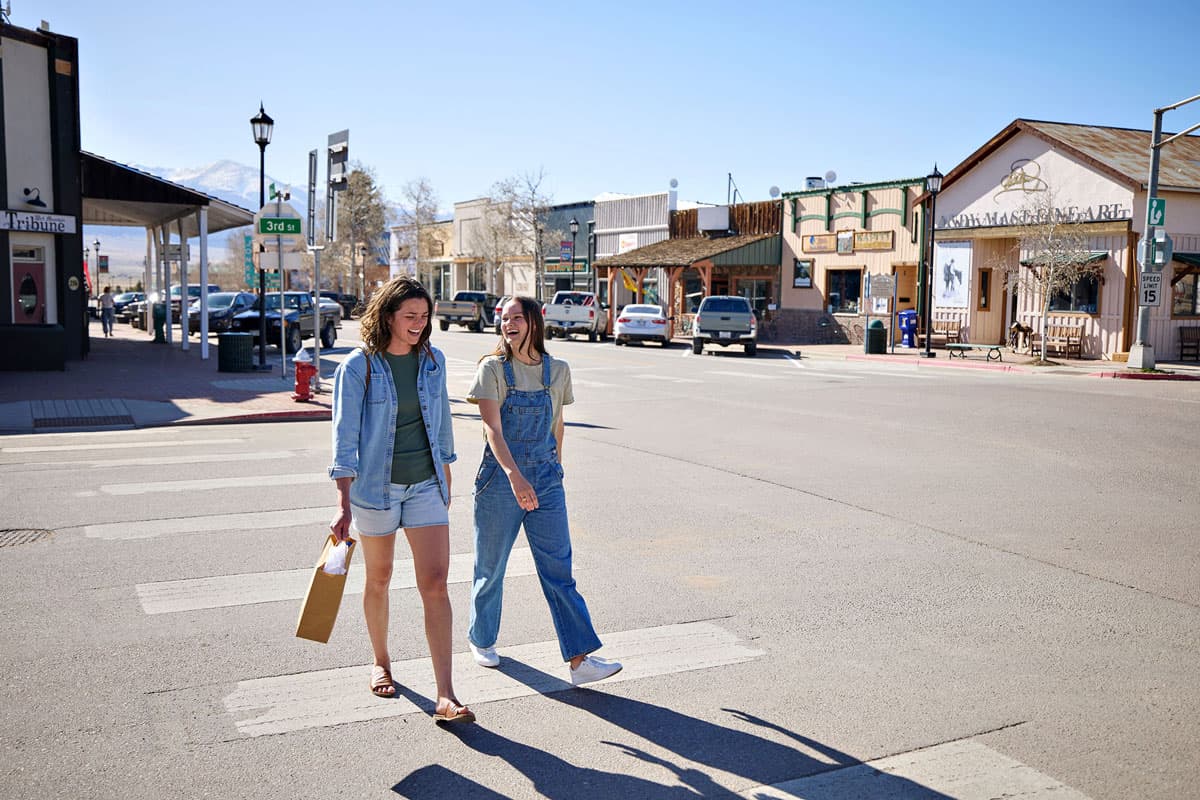 Two girlfriends crossing a road in downtown Westcliffe with a shopping bag in hand and stores behind them.