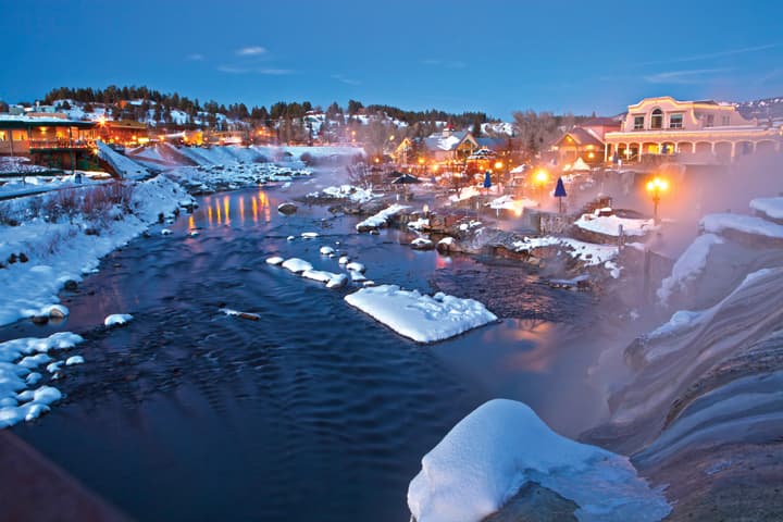 Warm lights glow over a steaming hot spring surrounded by snow in Pagosa Springs