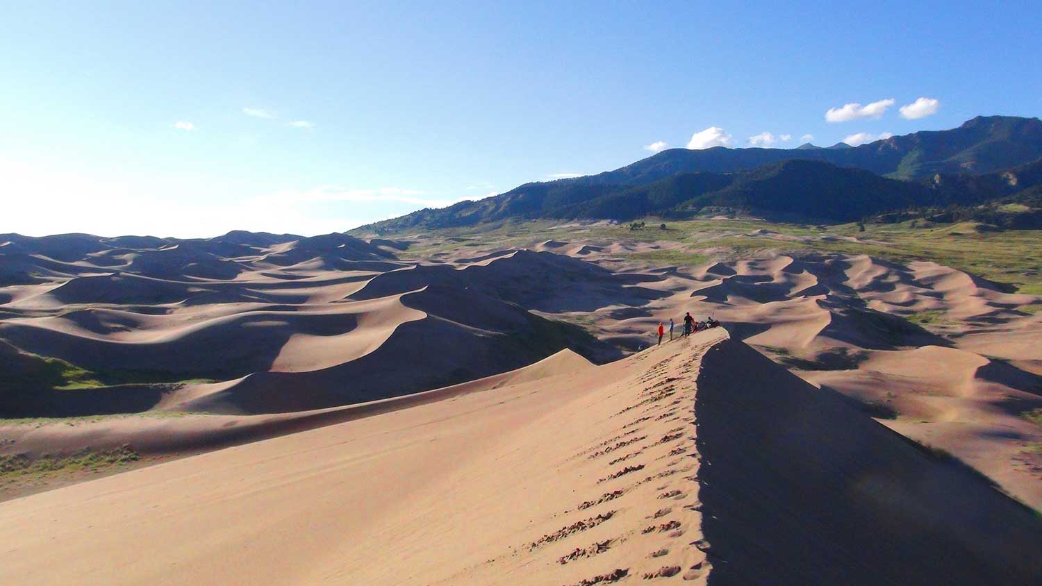 Tracks at the top of a large sand dune go off into the distance to where a group of people are. Behind them loom the Colorado Rockies.
