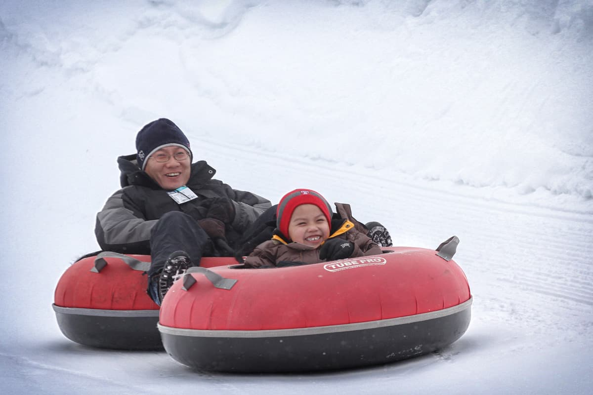 A parent and child flash big, happy smiles as they whiz down a slick, snowy slope in red and black inner tubes at Copper Mountain in Colorado.