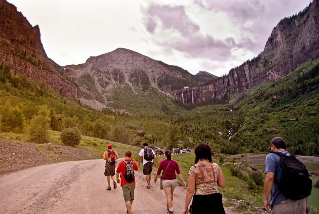 A group of six hikers walk along a wide dirt trail toward the cliffs of Telluride, where you can see a waterfall streaming down