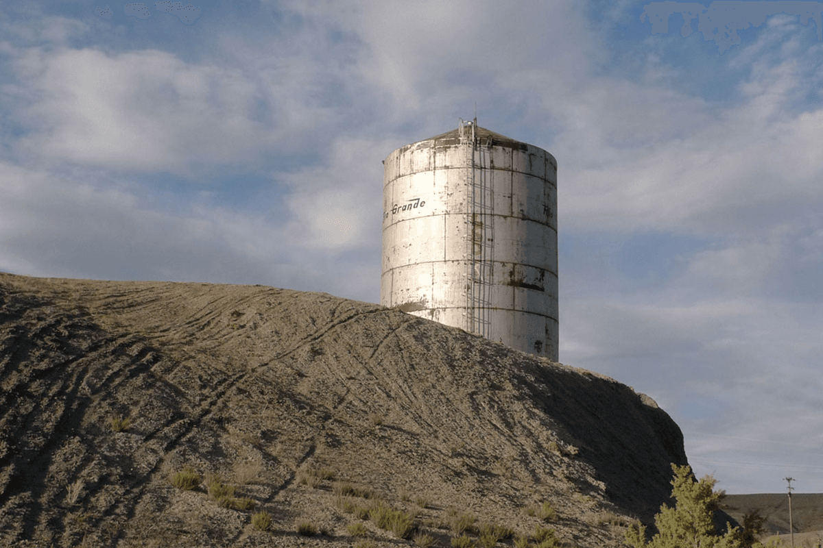 A metal silo sits behind a pile of grey rock against a hazy blue sky.