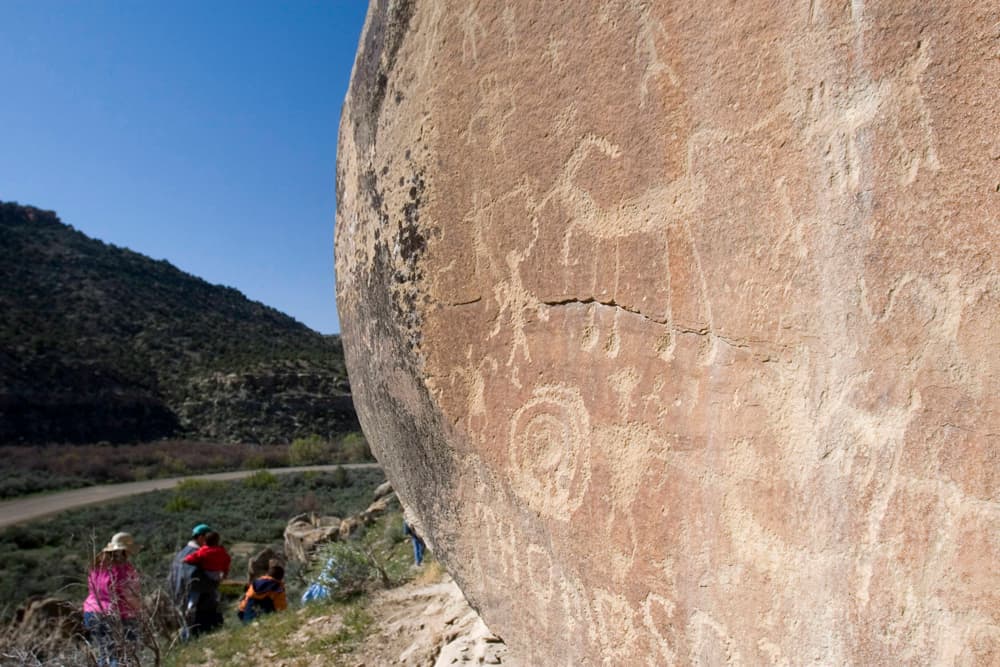 A sand-colored rock is covered in Petroglyphs and on the left side a family on a tour walks down the path during a blue-sky day in Ute Mountain Tribal Park.