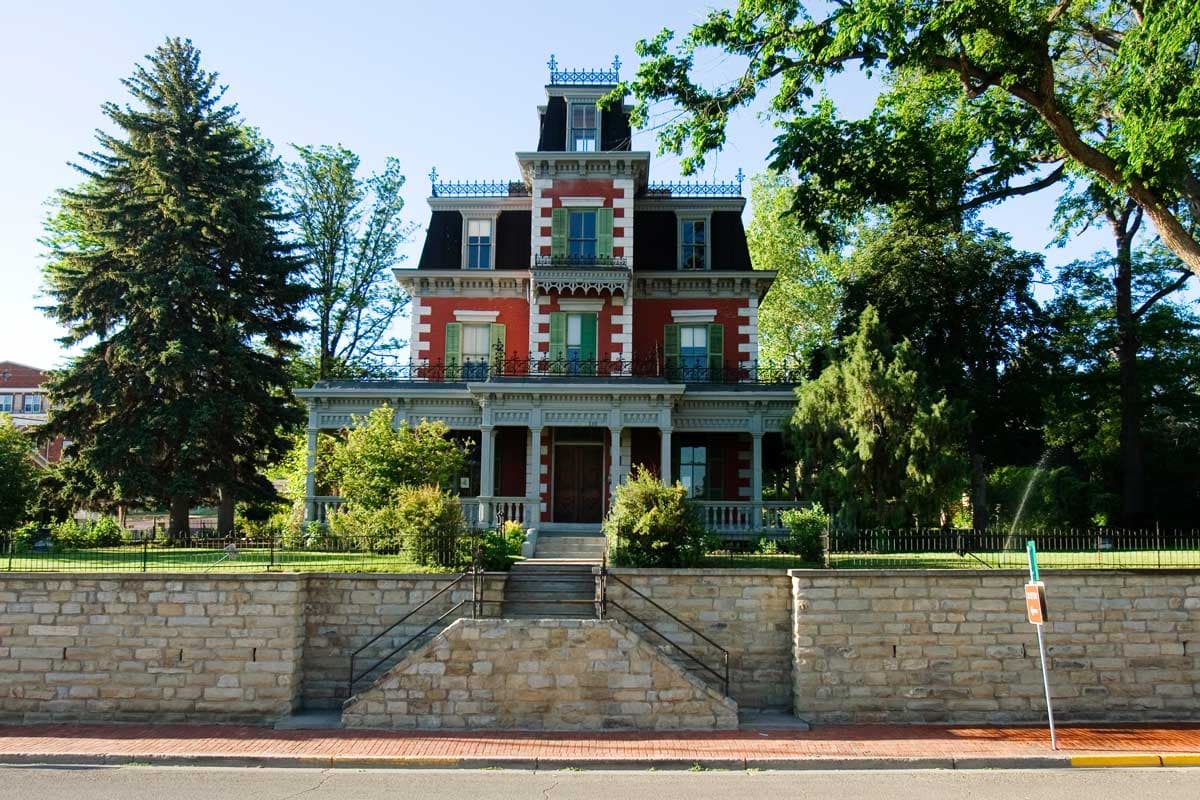 A three-story mansion pained red, with white brick accents, green shutters and a black mansard roof sits behind a brick wall and manicured lawn