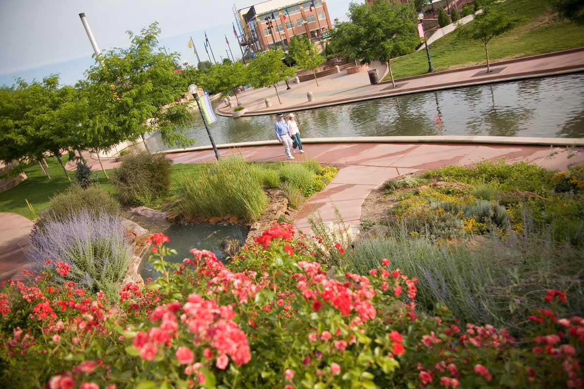 A couple walks along the Historic Arkansas Riverwalk with blooming flowers and green plants to the right.