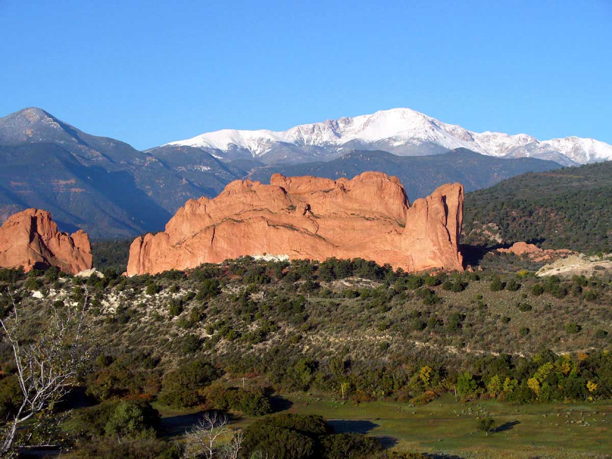 A giant red-rock geologic formation comes up from green grasses and scrubby bushes under the snow-covered Pikes Peak.