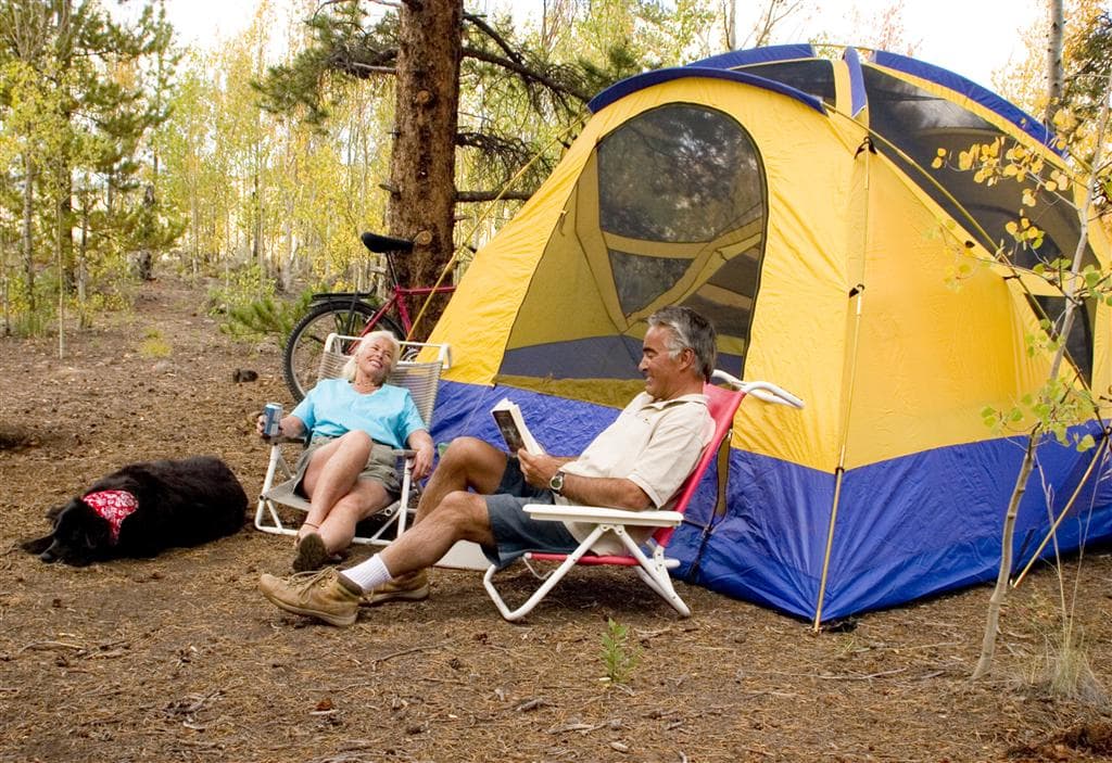 A man and woman sit in camp chairs in front of a yellow and blue tent in Crawford State Park. There's a dog sleeping to the left of the woman, who is wearing a blue shirt. There are skinny green trees in the background.