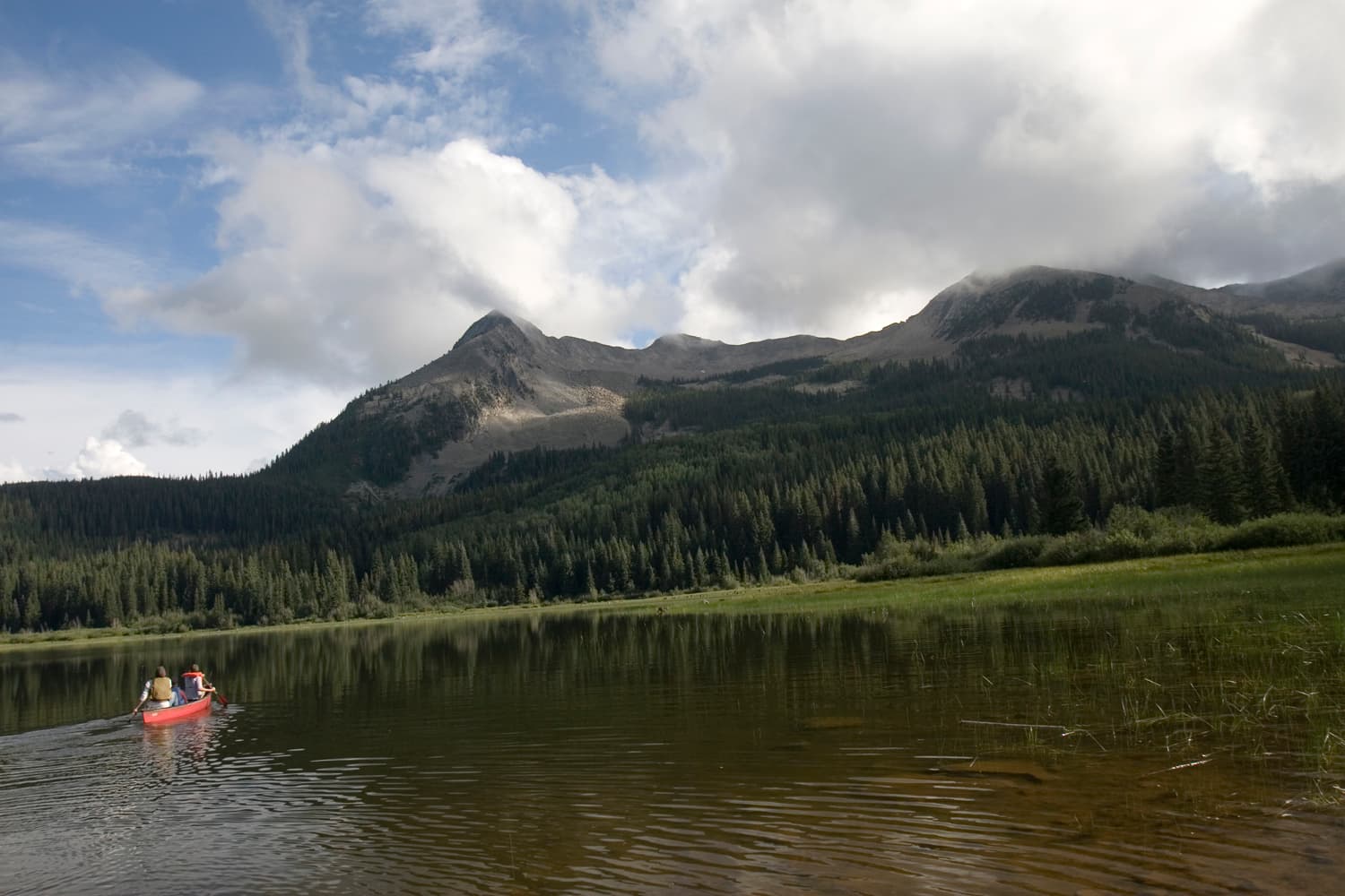A red canoe with two people in it sits in the middle of a calm lake with surrounded by evergreen trees. There's a stone-mountain peak in the distance.
