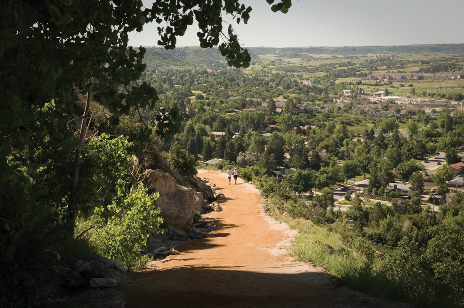 A red-clay path sits on the side of a mountain with greenery on both sides. In the distance, two people walk down. In the background the town of Castle Rock sits.