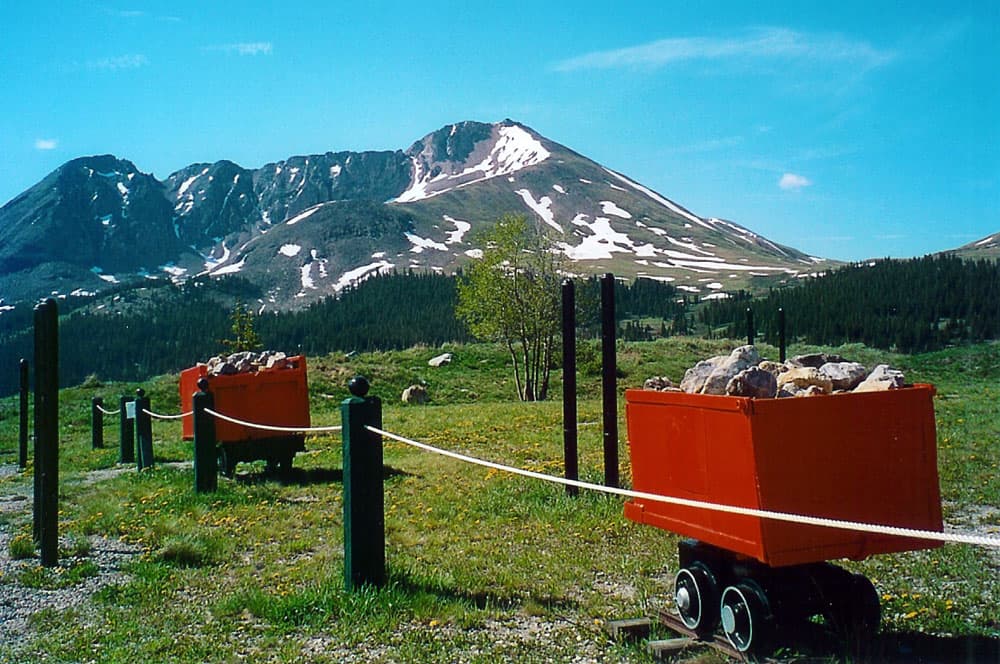On a summer's day two red mining cars sit with rocks behind a rope fence. There's green grass, evergreen trees and a snow-capped mountain sitting under a bright-blue sky.