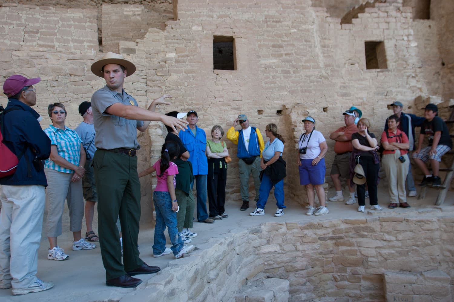 A ranger dressed in dark green pants, a khaki shirt and a wide-brimmed hat stands in front of an open pit and speaks to a tour group at Mesa Verde National Park.
