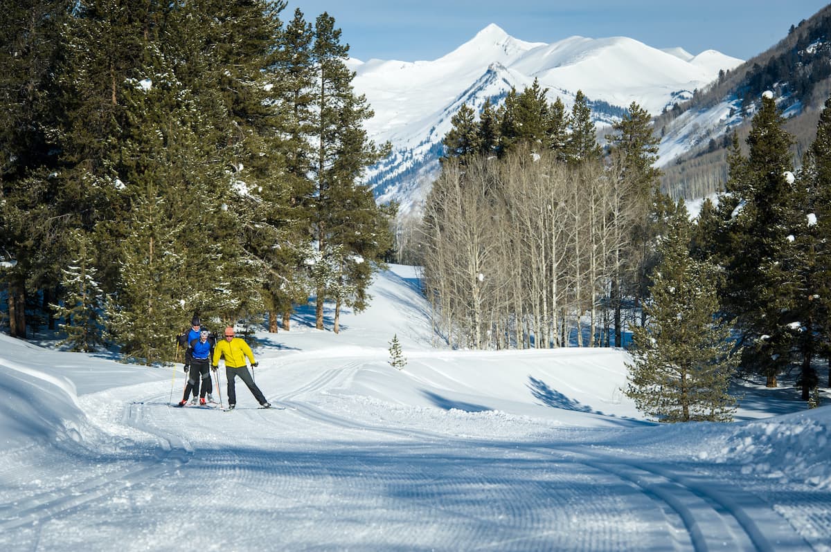 Three cross-country skiers travel across a groomed track with mountains covered in deep powder behind them