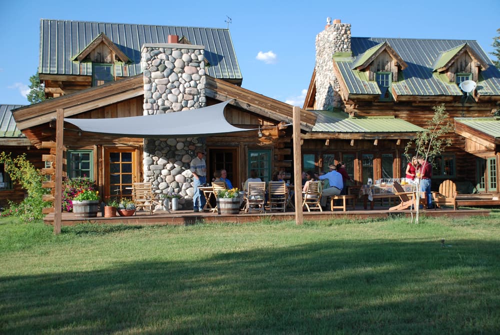 Two log cabins with stone fireplaces sit on green grass under a blue sky in the summer. People are sitting on the porch chatting.