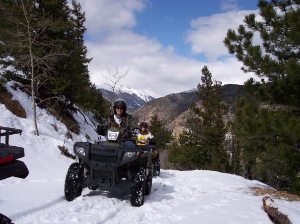 Two people sit atop two ATVs in a row on a snowy path with evergreen trees on both sides. The path is on the side of a mountain and there is a blue sky with white clouds above.