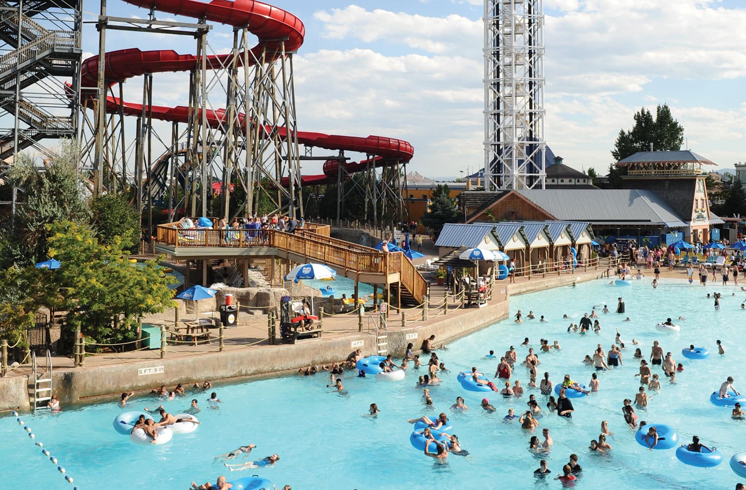 Hot vacationers swim and float on thick inner tubes in a large pool in Denver Colorado. To the side is a red, multi-story waterslide that snakes off down below a stand of trees.