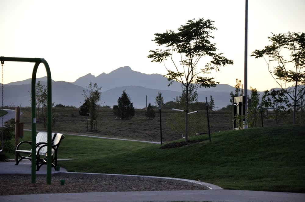 A sunset view of the Front Range mountains from a park in Erie. Recently planted trees sit near a bench with a golden sky.