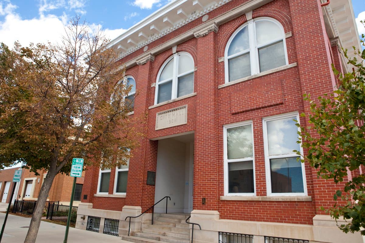 A two-story red-brick building sits at an angle with rectangular windows on the first floor and arched windows on the second floor. There are green trees at the street level.