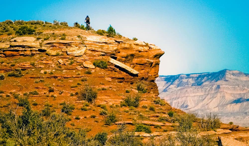 Way atop a giant orange-colored hill with splotches of sagebrush, we see a wee mountain biker making their way along a trail