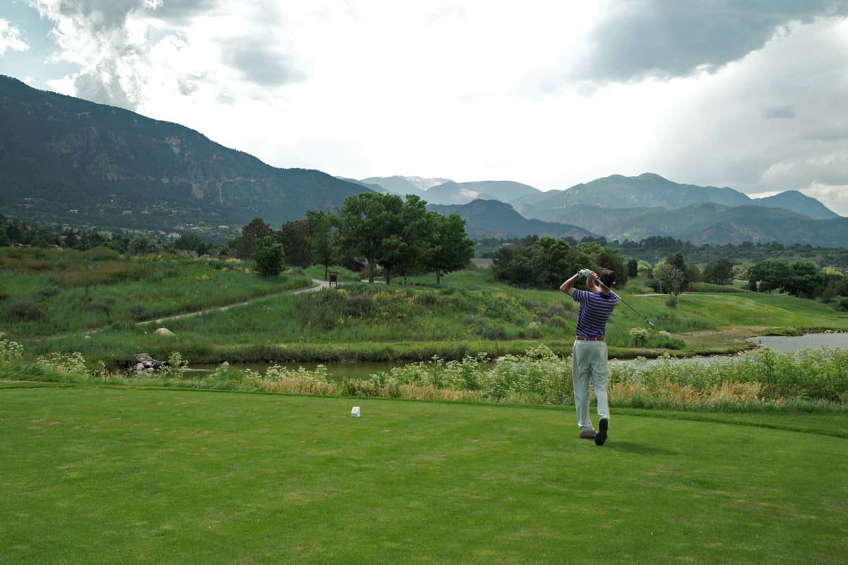 A person in a purple-striped shirt and khakis pauses at the end of their golf swing on a putting green in Colorado to see where their golf ball lands.