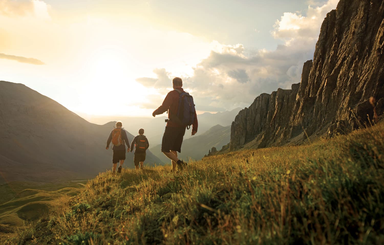 Hikers cross a mountain path as the sun glows over the horizon