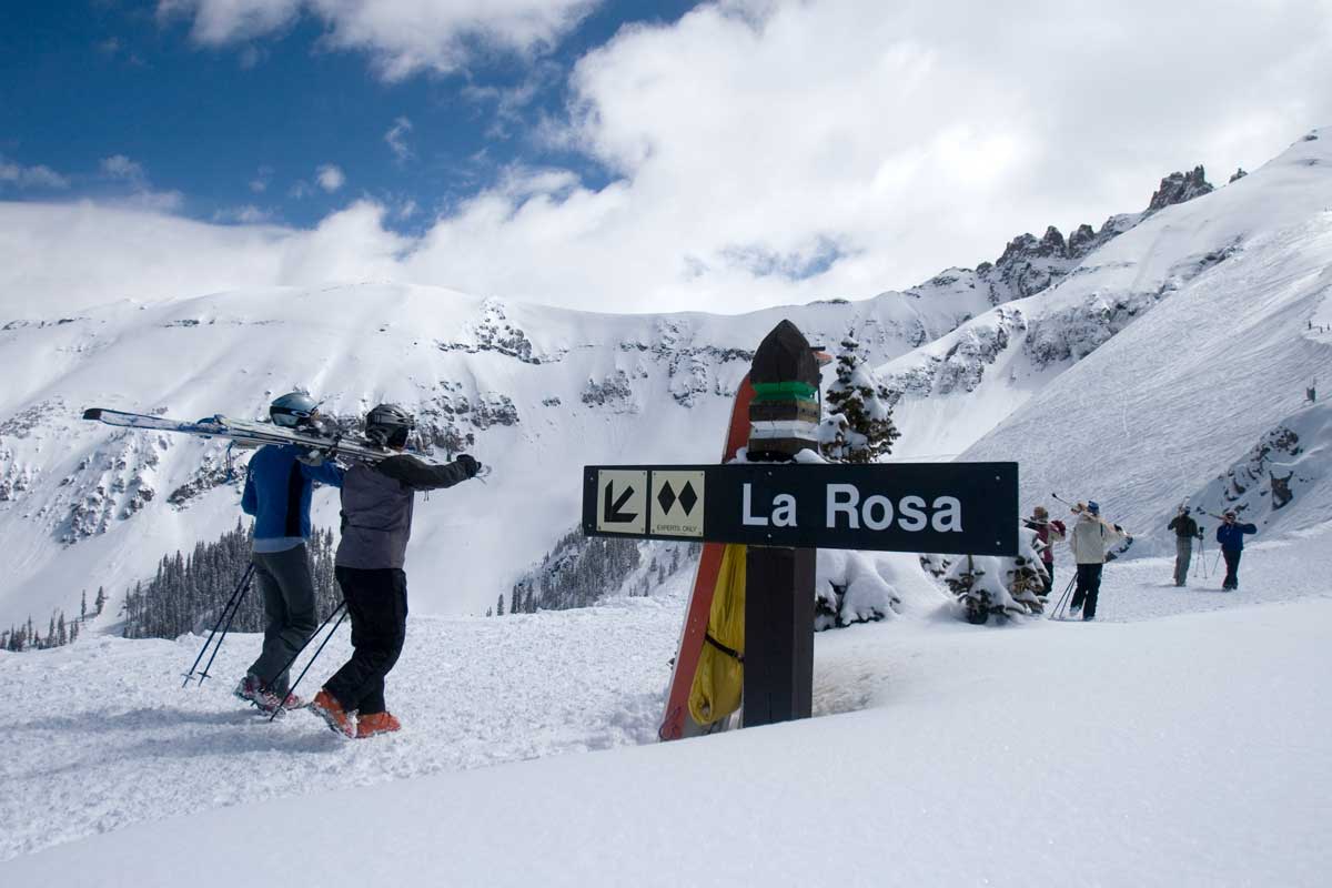 A winter's day skiing in Telluride with two skiers walking through the snow with their skis on their shoulders surrounded by snow-covered mountain peaks. A sign in the middle of the image says "La Rosa" in white lettering.