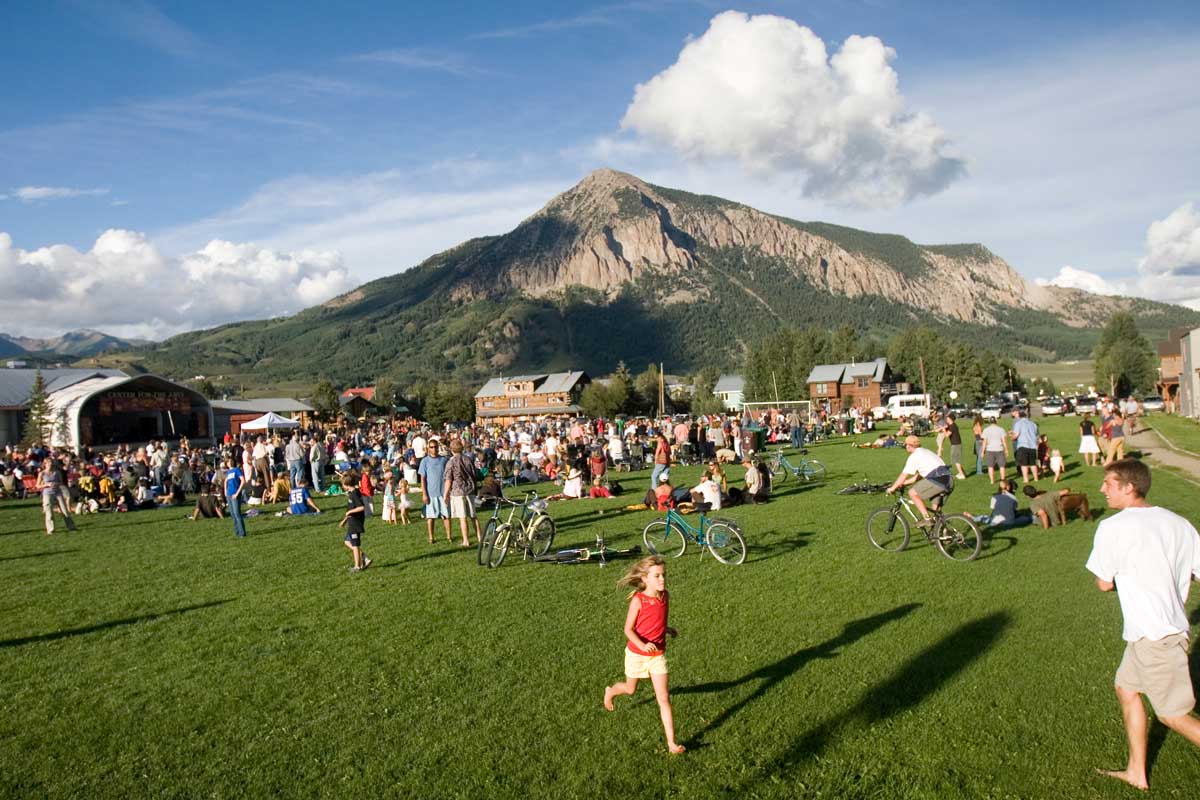 A summer concert in Crested Butte is taking place under a blue sky with cotton ball clouds. The grass is bright-green, there are bikes parked and children are running around with a rocky mountain peak in the background.