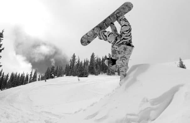 Snowboarder flipping mid-air above a snowy mound with trees in the distance at Monarch Mountain, Colorado.