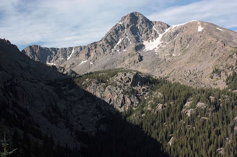 The rocky peak of Mount of the Holy Cross sits with snow nestled in a nook. Beneath it there are rocky outcroppings with evergreen trees.