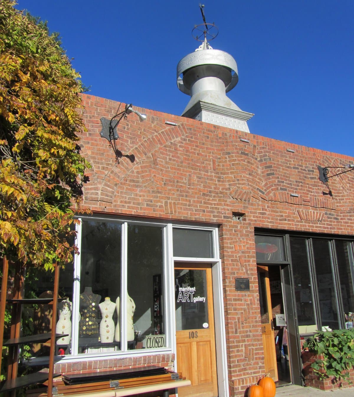 A red-brick building with a door that says "Manifest Art Gallery" sits under a blue sky.