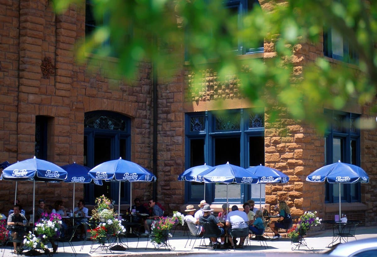 A red-stone historic building is shot through a green tree with seven Pepsi-Cola umbrellas with people sitting under them eating.