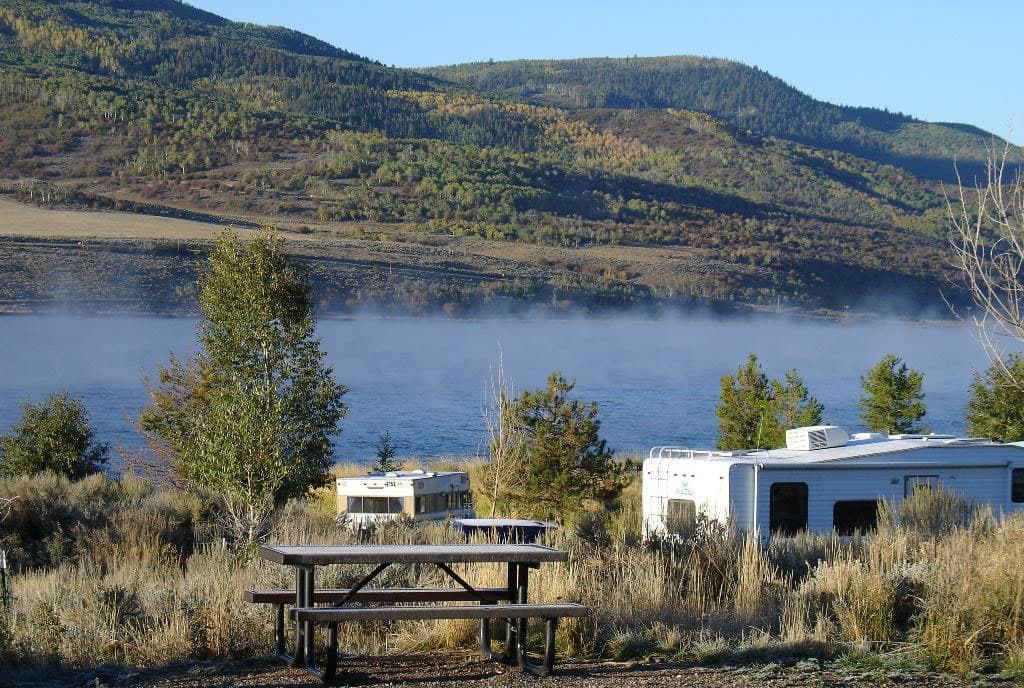 Two white RVs camp lakeside at Stagecoach State Park . There's a picnic bench in the foreground and a lake in the background with rolling, green-covered hills.