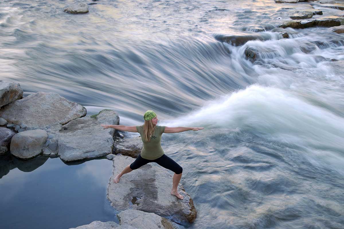 A person in yoga pants and t-shirt stands on flat rock and holds a Warrior Two position next to the San Juan River in Pagosa Springs, Colorado.
