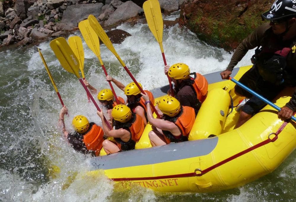 Six rafters wearing yellow plastic helmets with orange life jackets sit in a bright-yellow inflatable raft and tumble nose first into splashing whitewater.