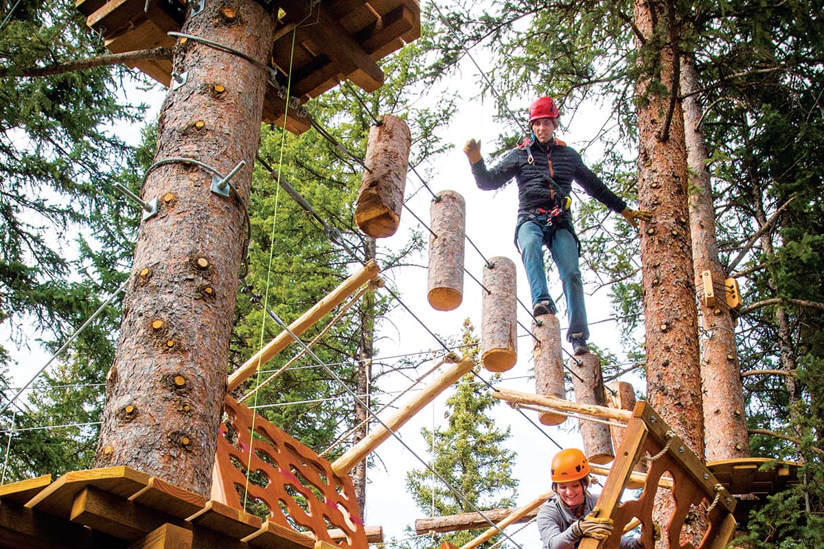 An adult wears a red helmet and is strapped into a safety harness as they explore a treetop playground and obstacle course among the pines at Lost Forest in Aspen, Colorado.