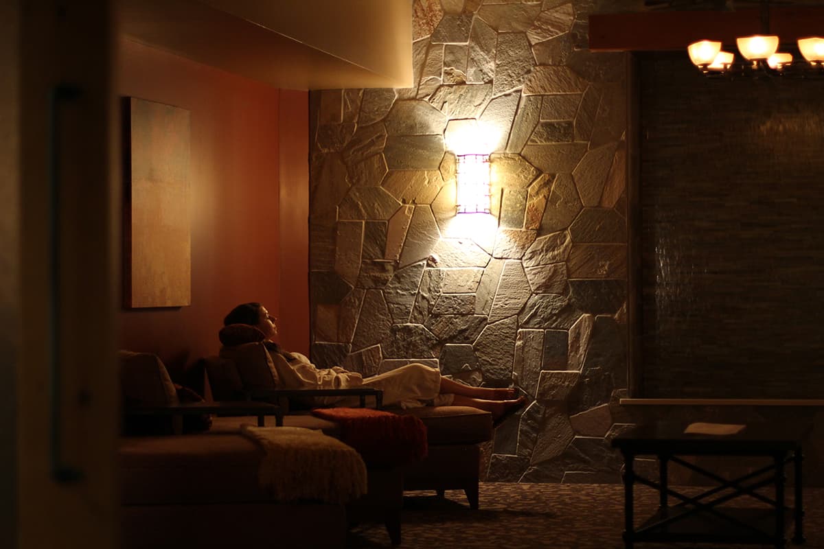 A light on a gray-stone wall creates a tranquil ambiance for a person reclining on a cushioned chaise in a white bathrobe at a spa in Colorado.