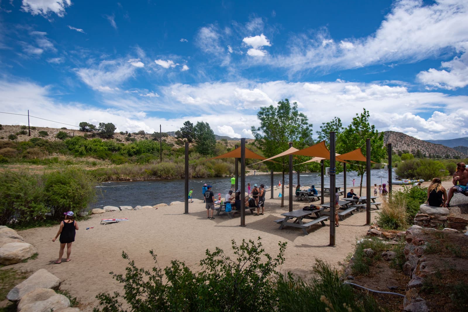 Alongside a calm river, several people eat lunch at picnic tables under yellow shades