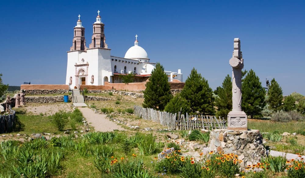 On a hill above a green-grass field with colorful wildflowers sits a bright-white church with two towers and a dome. The sky is blue, there are evergreen trees in the middle of the photo and a path leads through the grass to the Shrine of the Stations of the Cross building.