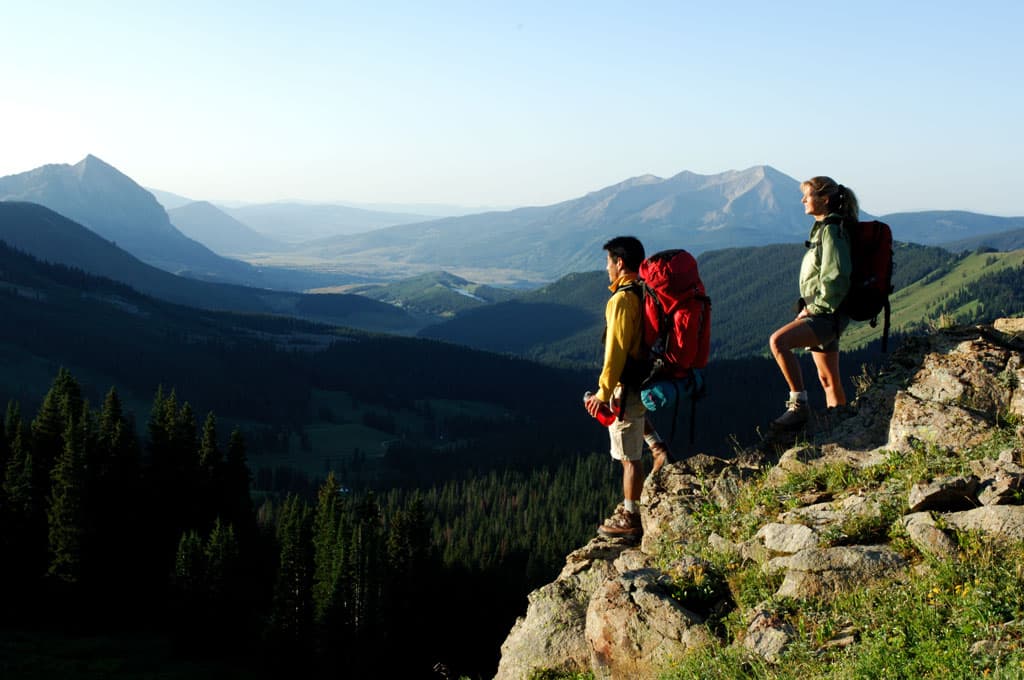Two people wearing big hiking backpacks stand at a cliff's edge and look across a canyon to blue peaks shaded by fog.