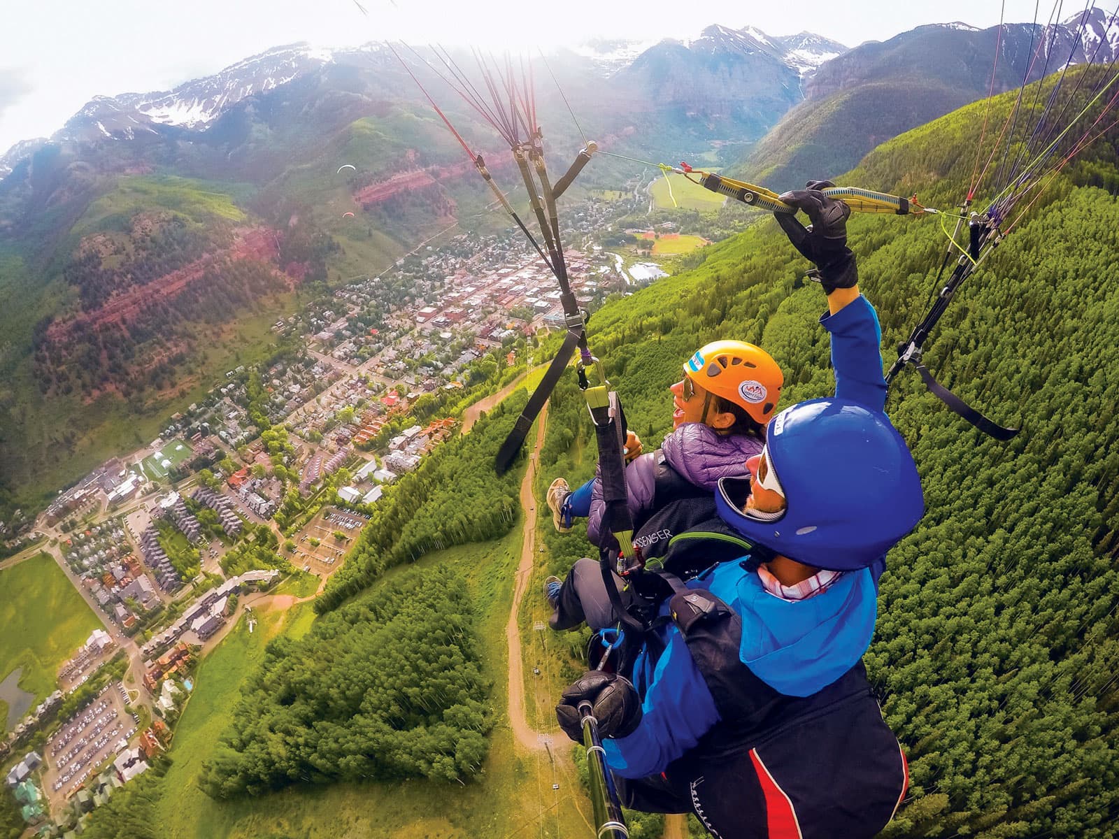 An eagle-eye view shows to people smiling and laughing as they paraglide together over a small town in Colorado nestled in a valley between two mountainside.