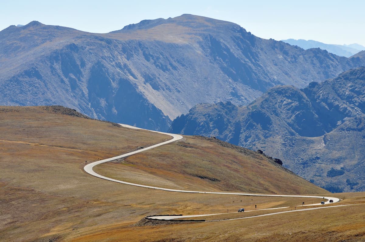 A handful of cars drive along the serpentine Trail Ridge Road in Rocky Mountain National Park in Colorado. In the background rugged mountain ridges seem to touch the sky.